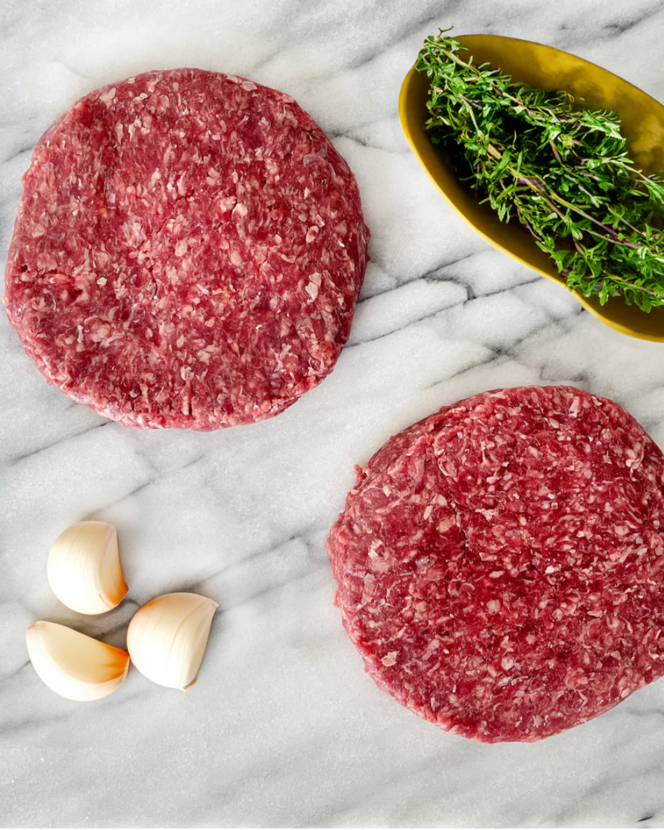 Two raw hamburger patties on a marble surface with garlic cloves and thyme.