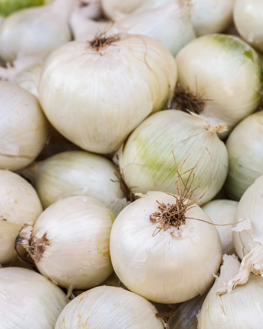 A close-up pile of white onions with dry brown tops, filling the frame.